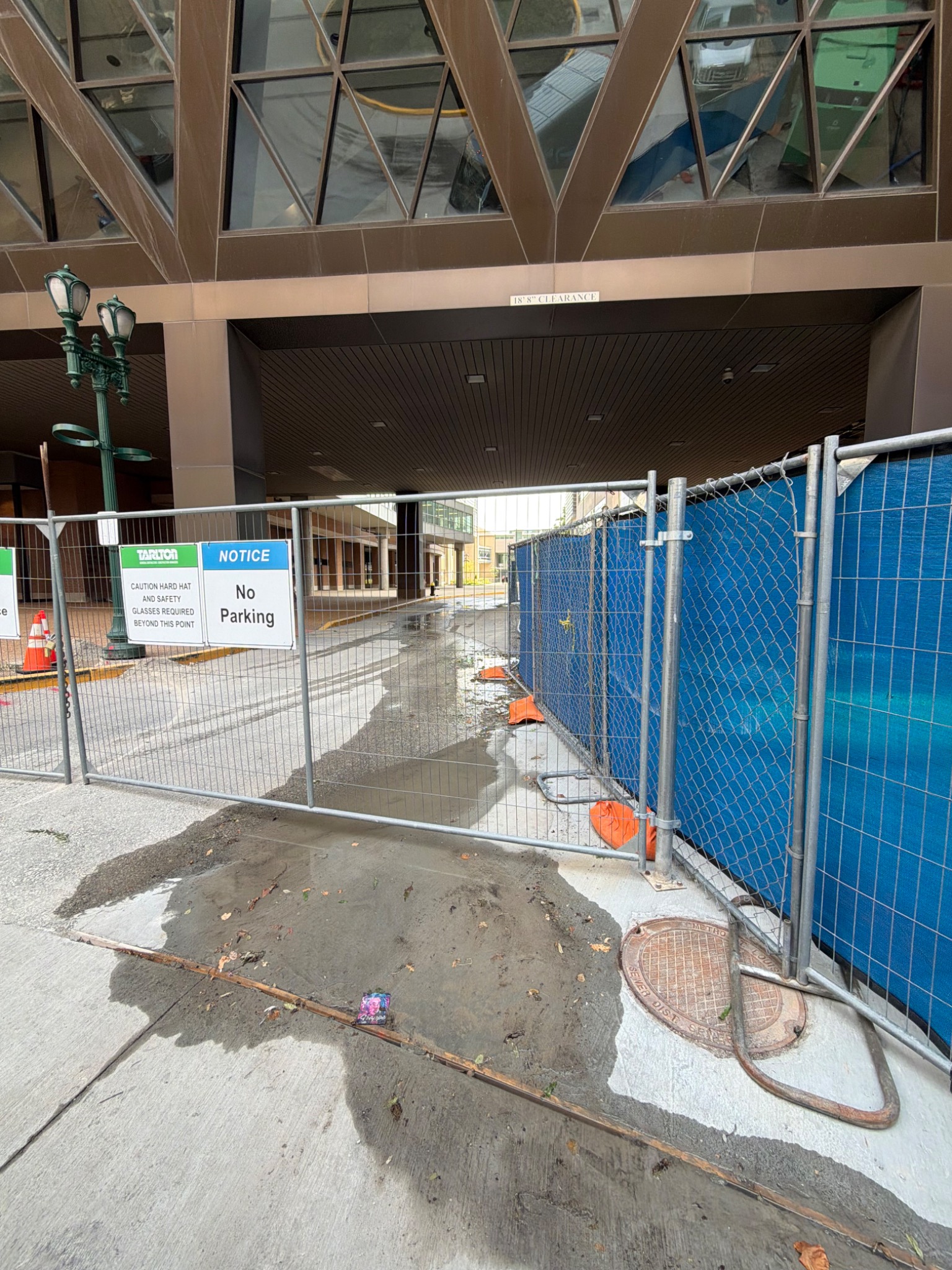 Construction fencing closes a driveway beneath a glass walkway between city buildings.